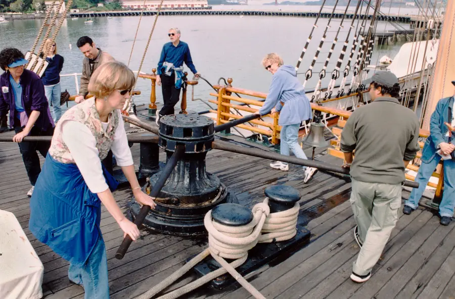 Visitors in casual clothes pushing wooden bars to turn an iron capstan on the deck of a sailing ship