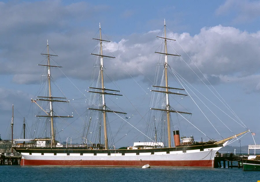 Three-masted square-rigged sailing ship with a black and white hull moored at a pier under a cloudy sky