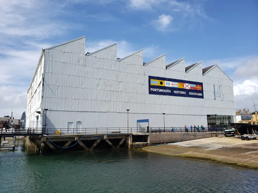 Large pale grey corrugated boathouse with saw-tooth roof gables standing on the water's edge at Portsmouth Historic Dockyard