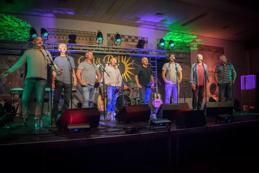 Eight men in striped and plain shirts singing at microphones on a lit concert stage