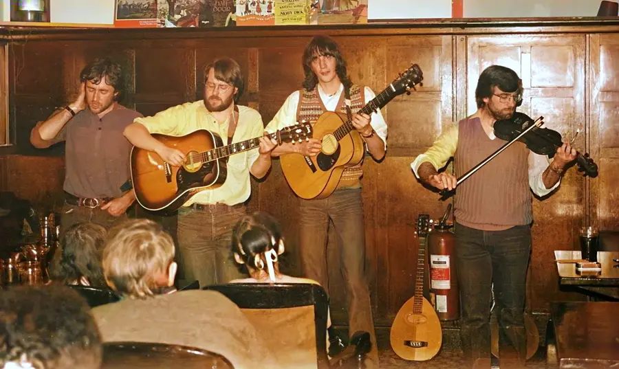 Four musicians with guitars and a fiddle playing to a seated audience in a wood-panelled folk club