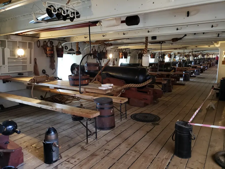 The gun deck of HMS Warrior with long mess tables, benches and black cannon between the gunports