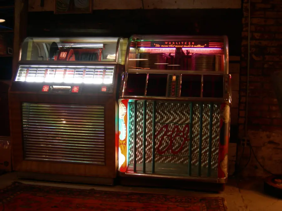 Two 1950s jukeboxes, a Seeburg and a Wurlitzer, glowing in a dark room