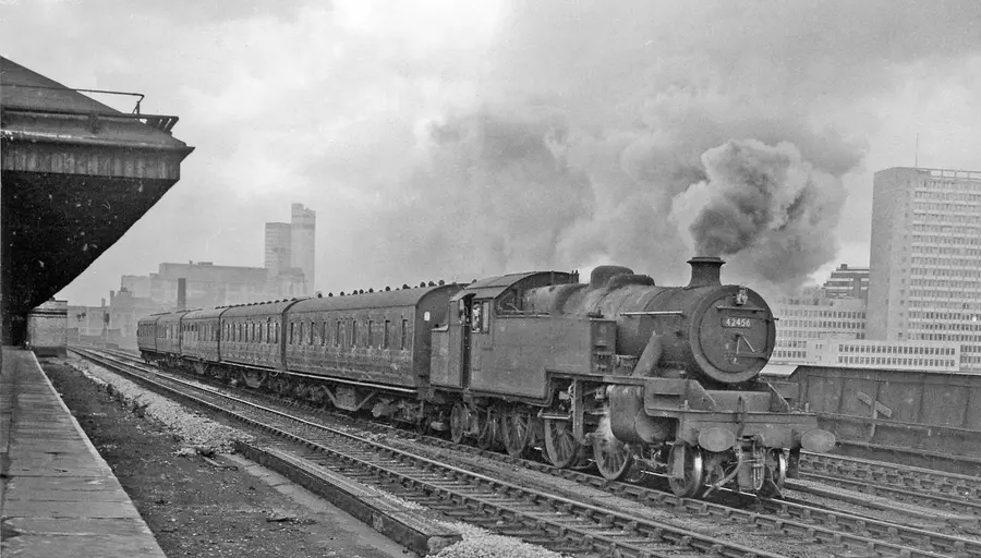 Black and white photograph of a steam locomotive pulling a passenger train past a station platform under heavy smoke