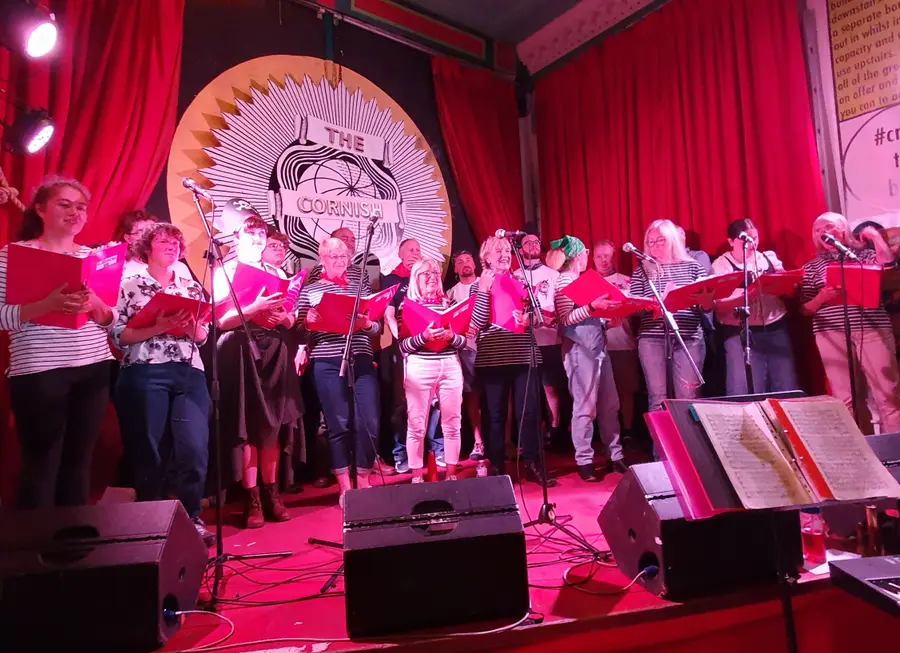 A shanty group in striped tops singing from red song folders on a stage under red lights