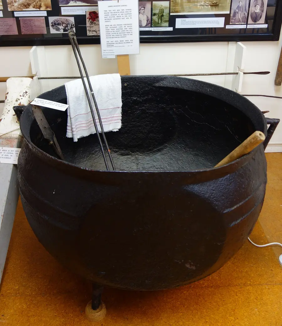 Large black cast-iron try pot on a museum floor with harpoons and labels behind it